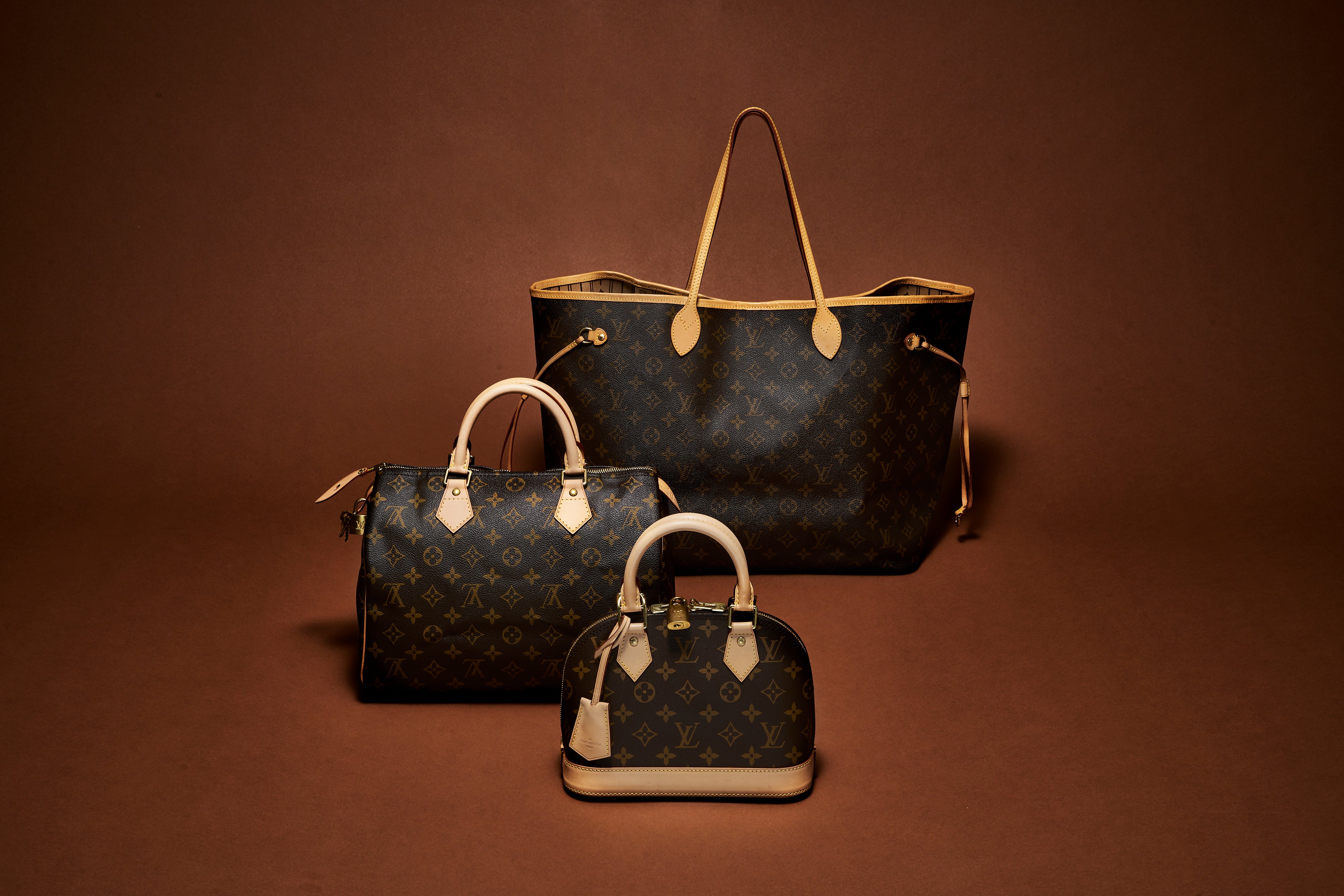 Three Louis Vuitton monogram handbags in different sizes arranged on a warm brown studio background: a large tote bag, a medium duffel-style bag, and a small top-handle satchel.