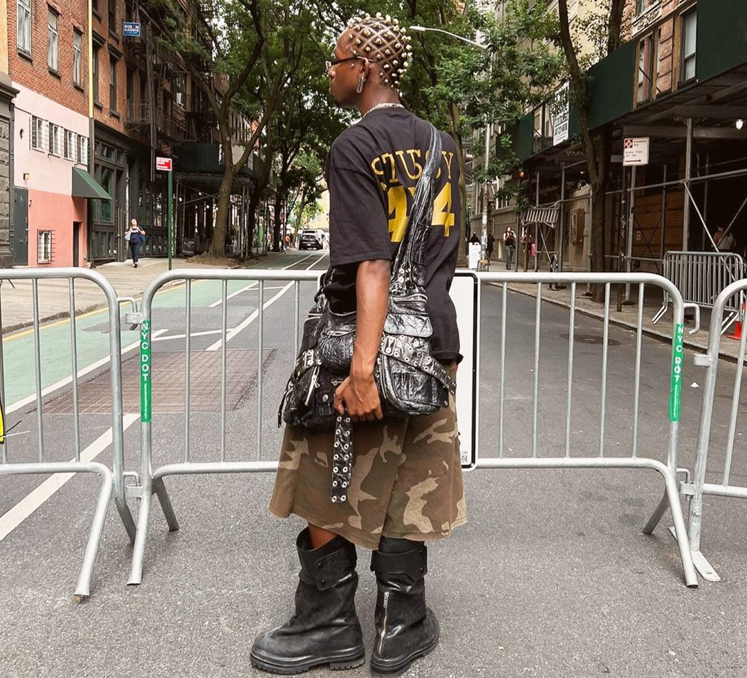 a man wearing black t-shirt, camo shorts, black leather boots and black leather crossbody bag