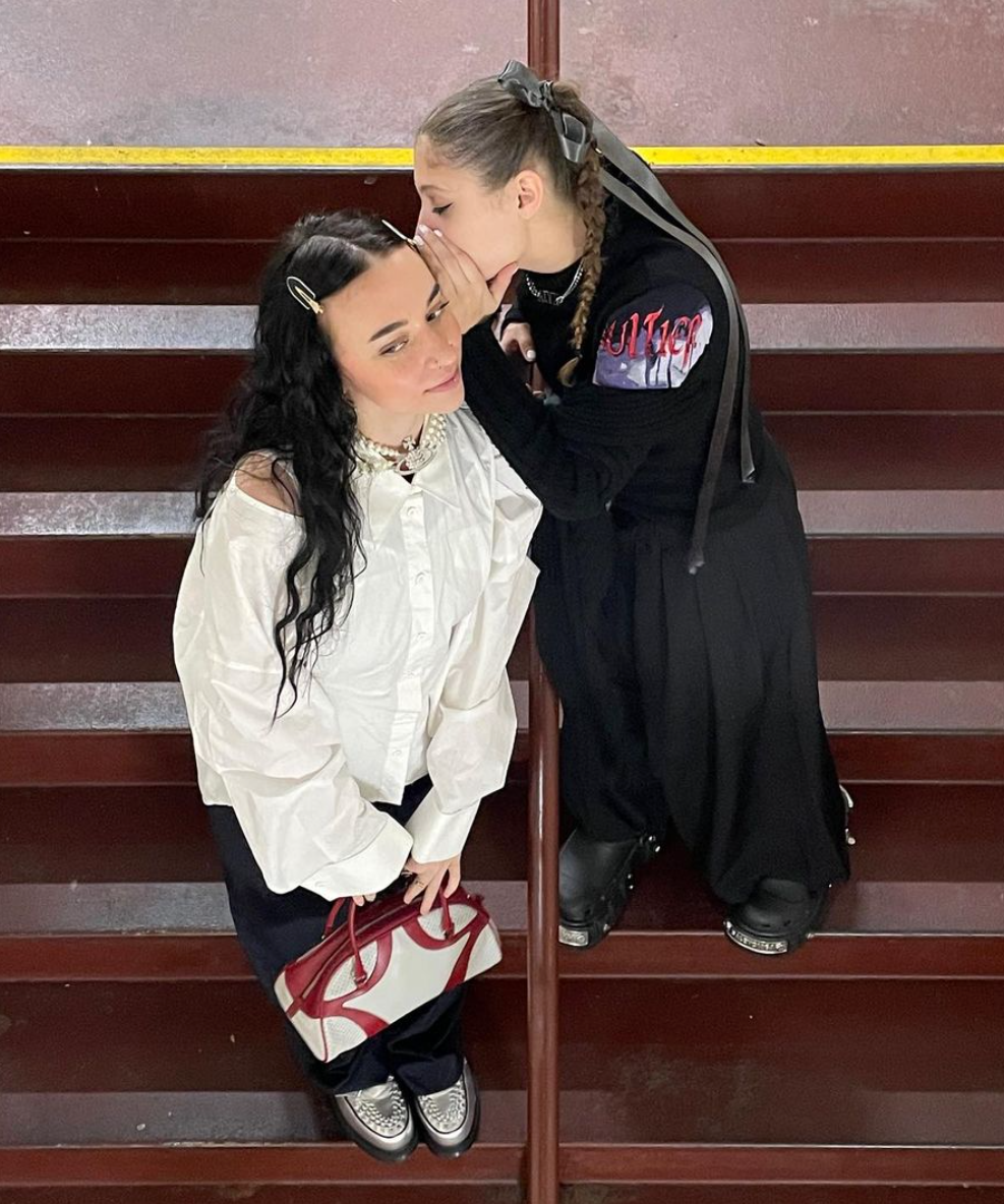 Two women posing on dark wooden stairs. One woman wears a white off-the-shoulder blouse, black skirt, and holds a white handbag with a red bow.
