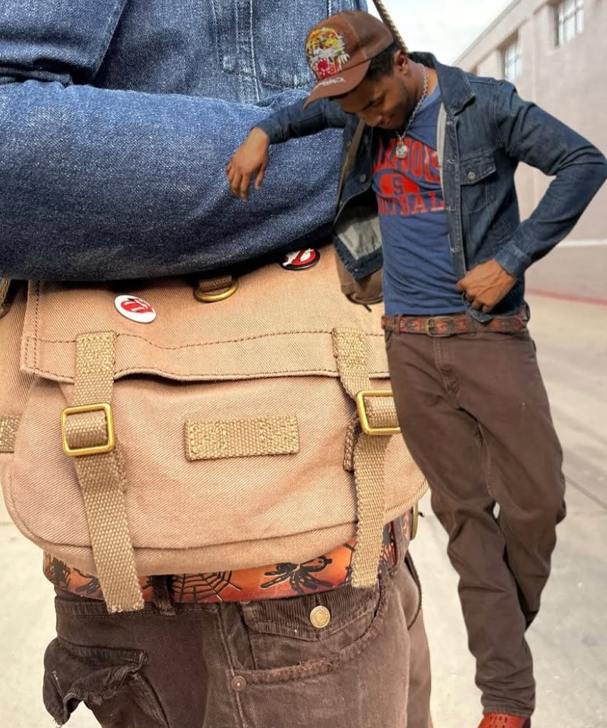A tan canvas messenger bag with gold-tone hardware and decorative pins, worn crossbody over a person's shoulder. A man in a denim jacket and blue vintage graphic shirt stands behind him in an outdoor urban setting.