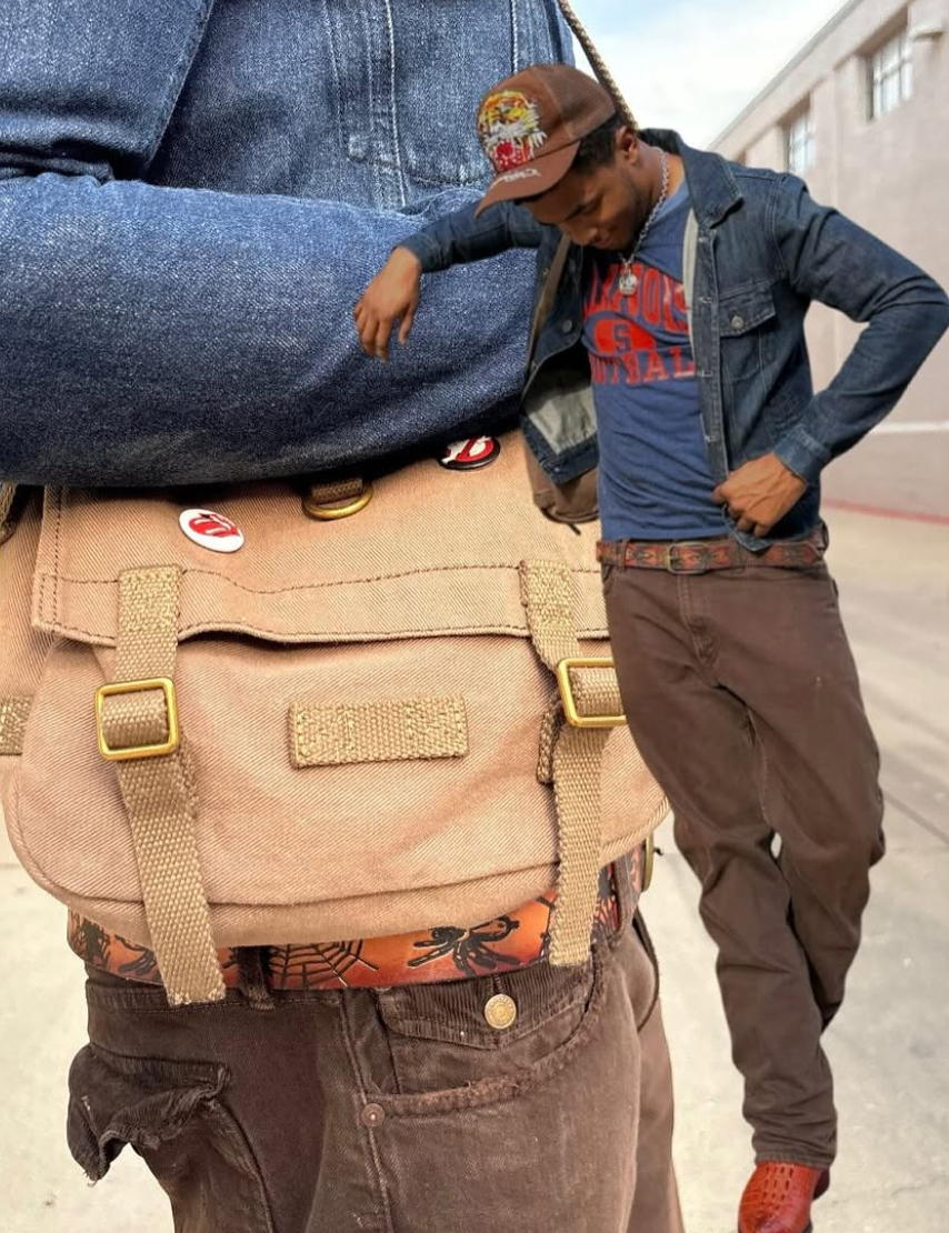 A tan canvas messenger bag with gold-tone hardware and decorative pins, worn crossbody over a person's shoulder. A man in a denim jacket and blue vintage graphic shirt stands behind him in an outdoor urban setting.
