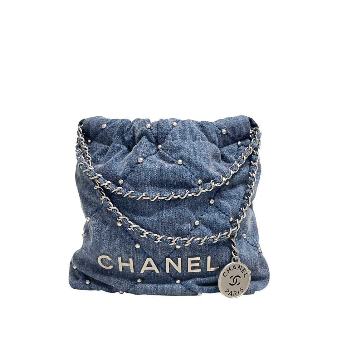 A Chanel denim shoulder bag with silver chain strap, rhinestone studs, and a silver-tone round charm engraved with the Chanel logo, shown on a plain white background.