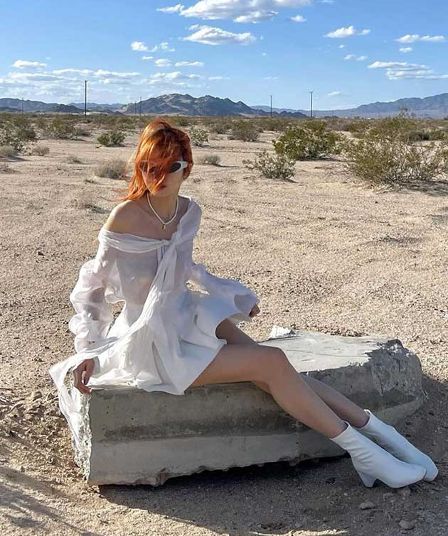 Person in a white dress sitting on a concrete block in a desert landscape with mountains in the background.