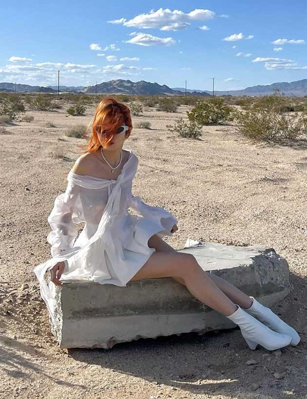 Person in a white dress sitting on a concrete block in a desert landscape with mountains in the background.