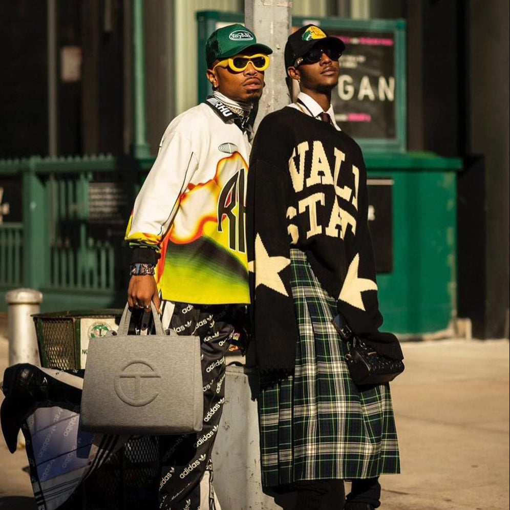 Two men standing on a city sidewalk wearing streetwear. One wears a white and neon yellow graphic sweatshirt, black pants with Adidas logos, a green cap, and sunglasses while holding a gray Adidas bag.