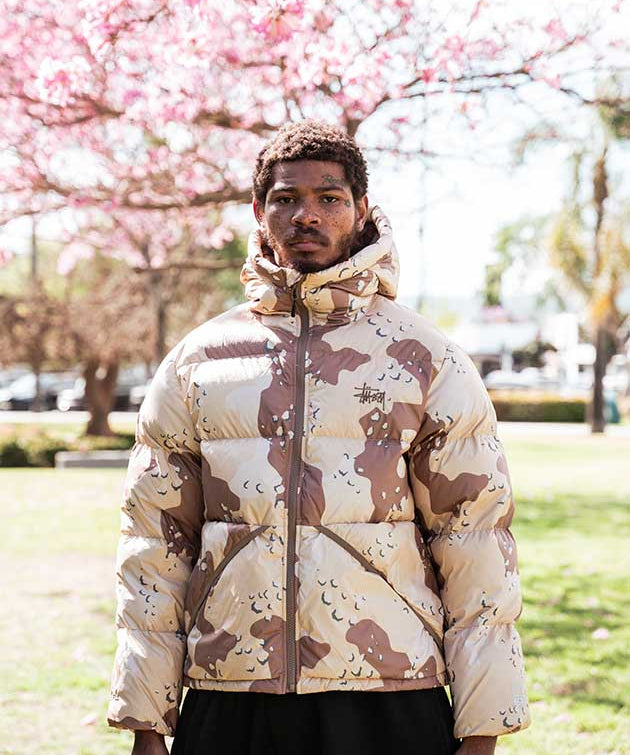 Person wearing a camouflage-patterned puffer jacket in a park with cherry blossoms.