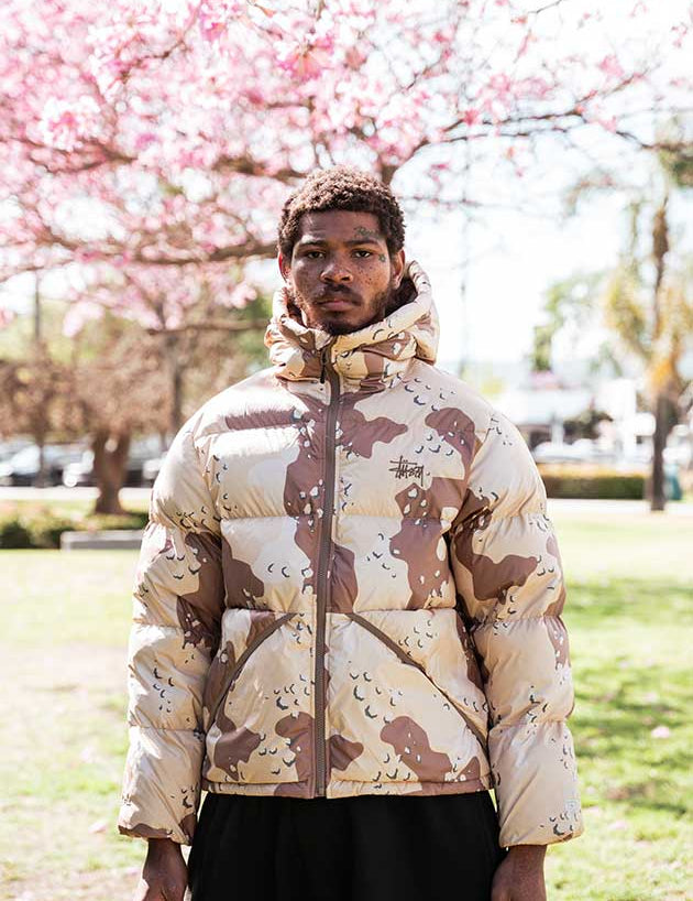 Person wearing a camouflage-patterned puffer jacket in a park with cherry blossoms.
