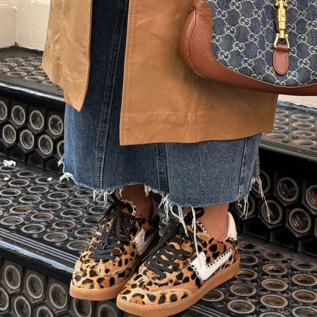 Person wearing leopard print sneakers with a brown jacket and denim skirt on a patterned floor.